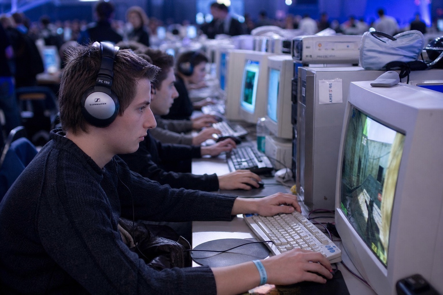 Salle de LAN party des années 2000 : longues rangées de PC et d'écrans CRT, joueurs casqués concentrés dans la lueur des écrans
